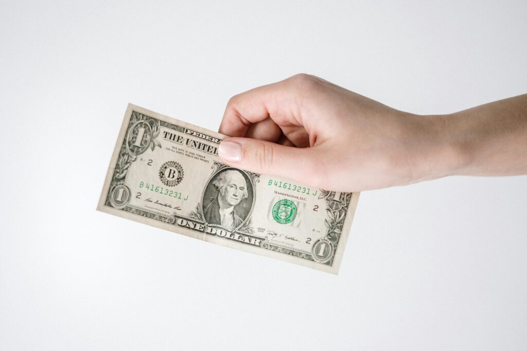 The image shows a hand holding a one-dollar bill against a plain white background. The hand is positioned from the right side of the frame, with fingers grasping the top edge of the bill. The dollar bill features the portrait of George Washington, as is typical for US one-dollar currency. The overall composition is simple and focused on the exchange of money.