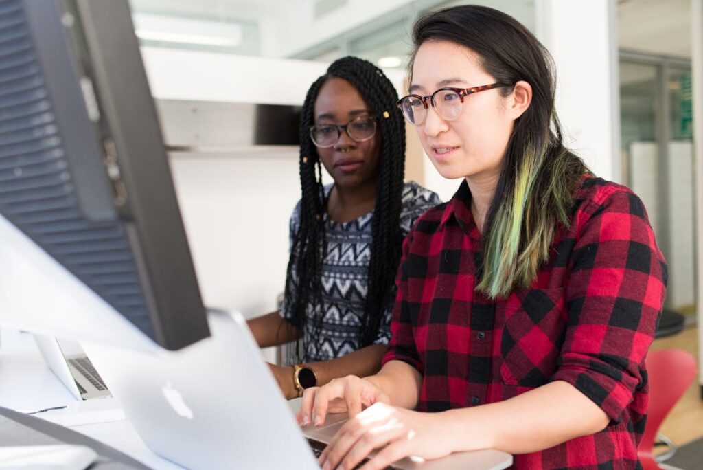 The image shows two women working together at a computer in a bright office setting. The woman in the foreground has long black hair with green highlights, wears glasses, and is dressed in a red and black plaid shirt. She is typing on a laptop. The woman in the background has long braided hair, wears glasses and a patterned shirt, and is looking at the computer screen. Both women appear focused on their work.