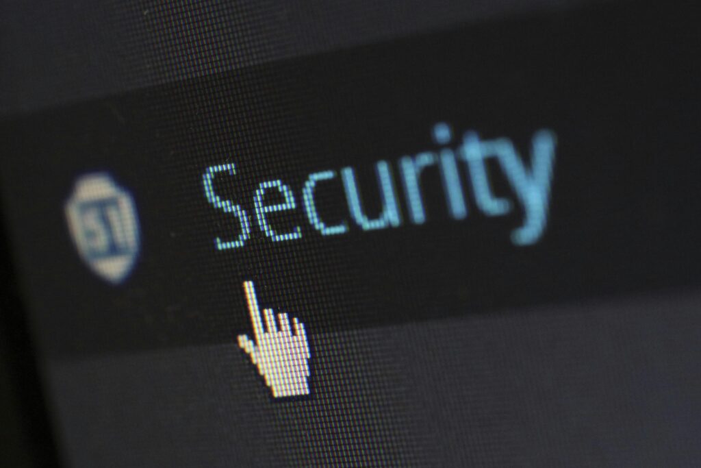 The image shows a close-up of a computer screen displaying the word "Security" with a hand-shaped cursor pointing at it. The screen has a pixelated look, and there is a shield icon next to the word "Security" on the left side. The overall impression is focused on cybersecurity or digital security.